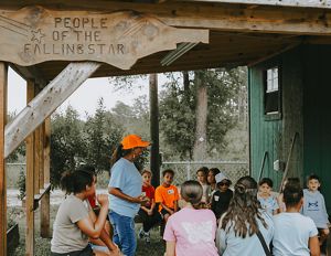 A woman in a bright orange hat speaks to a group of 14 youths.