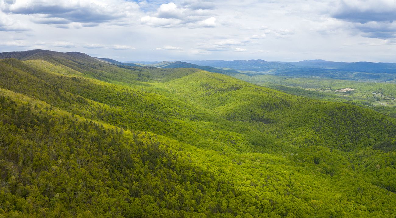 Drone view looking out over rolling mountain ridges and valleys. The mountain sides are thickly forested. Sunlight dapples over the trees. Small clearings and farms are visible in the distance.