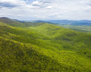 Drone view looking out over rolling mountain ridges and valleys. The mountain sides are thickly forested. Sunlight dapples over the trees. Small clearings and farms are visible in the distance.