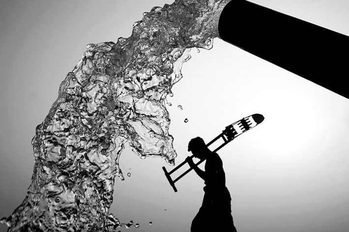 A black and white view of water pouring from a spout and the silhouette of a person walking and carrying a large tool.