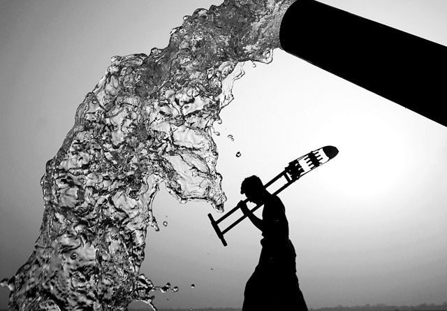 A black and white view of water pouring from a spout and the silhouette of a person walking and carrying a large tool.