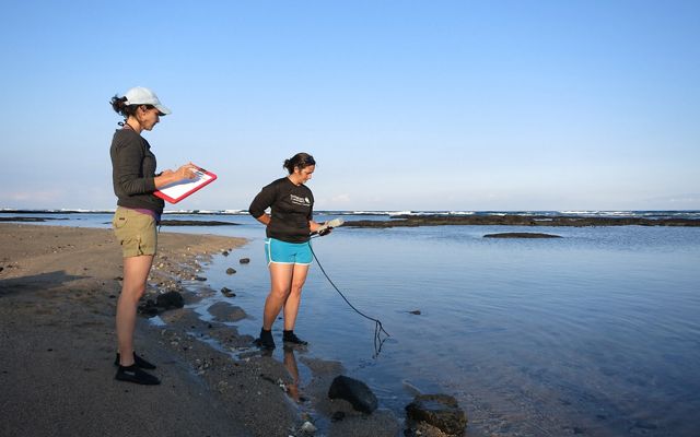 Two researchers stand on a beach with clipboards and a water monitoring instrument.