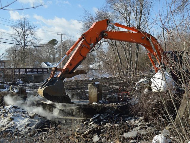An orange excavator arm poised above the concrete remains of the West Britannia Dam on a winter day.