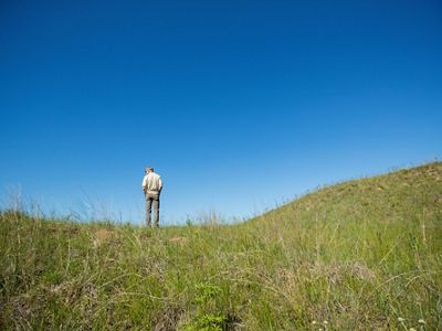 A man stands atop a sandy, grassy hill.