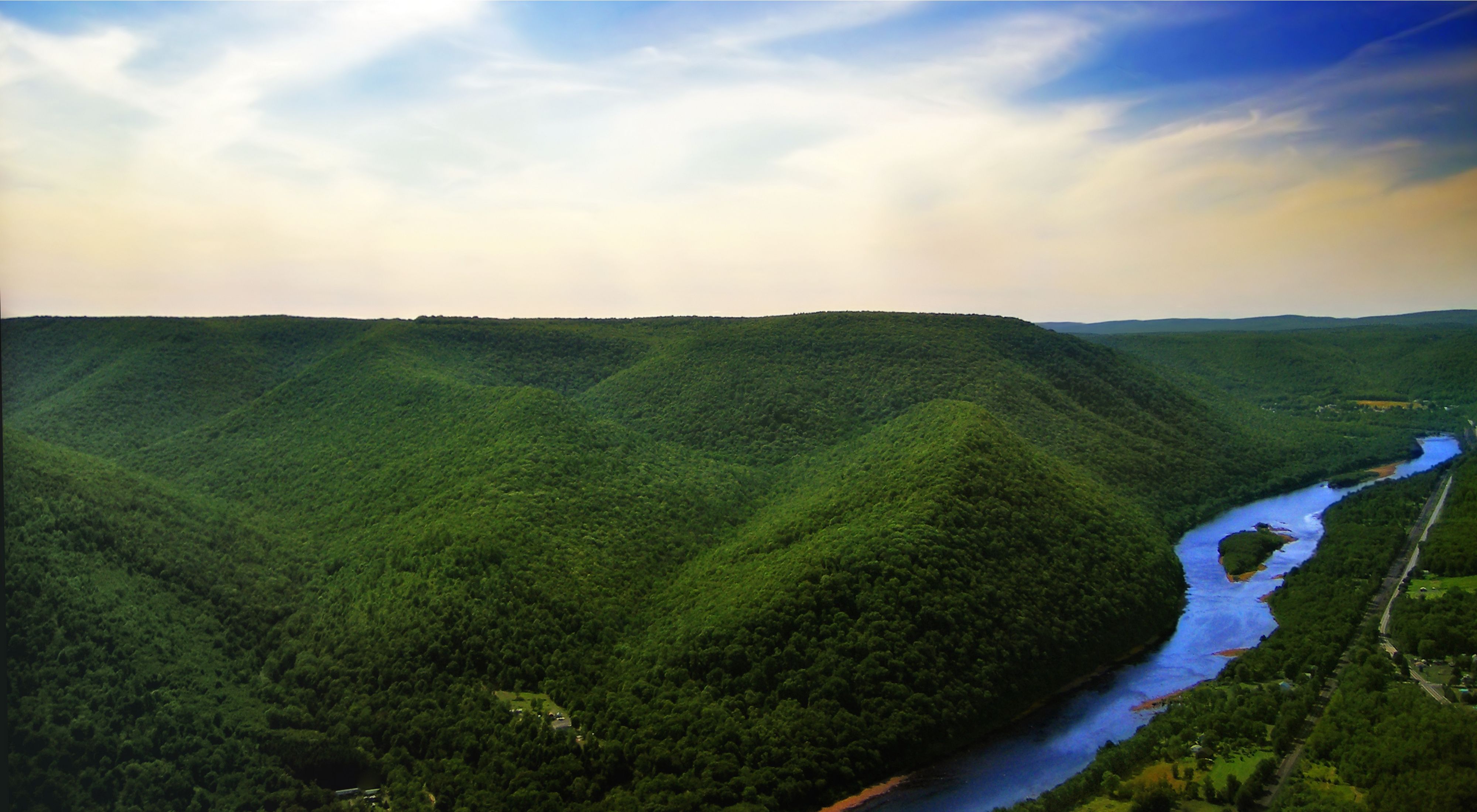 A vista view featuring rolling green hills and a bright blue sky with white clouds. A river gently flows to the right of the hills.