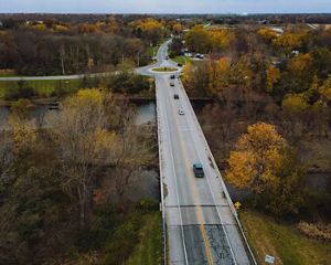 The White River in east central Indiana. Cars drive on a two-lane bridge that spans a narrow river. Buildings peek from beneath the trees that grow along the river bank.