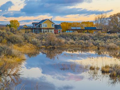 A yellow and red building reflected in a silver body of water with trees along its banks.