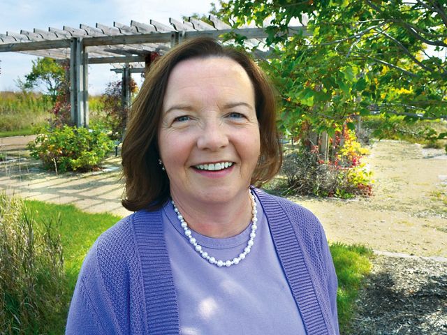 Woman wearing a blue shirt and sweater and a string of pearls stands in front of a trellis with flowers in a patio garden and smiles at the camera.
