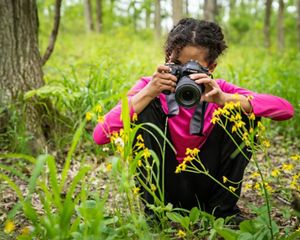A young girl sits on the ground to take a photo of wildflowers.