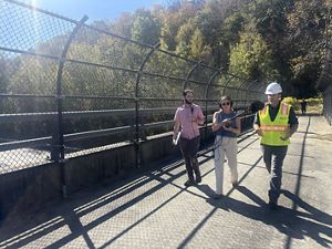 Two podcast producers, one holding a large fuzzy microphone, walk on a pedestrian overpass over the four lanes of the Massachusetts Turnpike, from forested area on one side to another. 