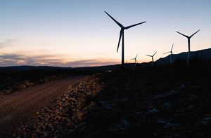Wind turbines stand on a hilly landscape against an orange sky.