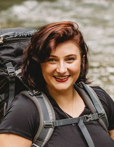 Headshot of Windy Daniels with a black shirt and a gray backpack with a creek in the background.