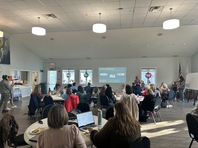 People sit at round tables in a community center with one person presenting in front of a screen.