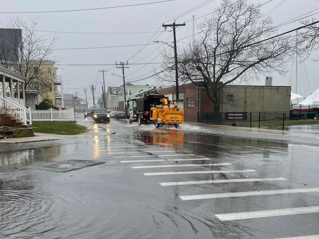 Flooded streets at an intersection with houses on the left side and a concrete building surrounded by a black chain link fence on the other side.