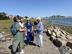 Four people stand on the rocky coastline of the southern edge of Winthrop, with a small harbor in the background. The two people on the left are holding microphones, recording the other two. 