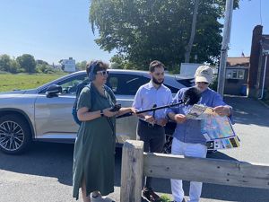 Two podcast producers, one holding a large fuzzy microphone, stand with Norman Hyatt, who holds a map of Winthrop. They're standing in the parking area of shoreline access in south Winthrop.