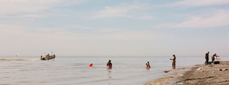 Several adults and children process fish on a beach in Bristol Bay, Alaska, while a small fishing boat motors away from the shore.