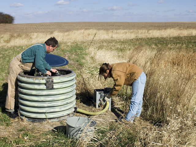 Two people work in a field. One person works in a large drum containing electrical equipment, and the other attaches a hose to a mechanical box.