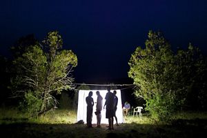 At nighttime, four people stand in front of a brightly lit sheet surrounded by tall trees.