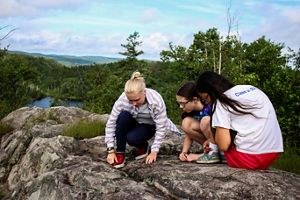 Three people crouch looking at a rocky outcropping above forests and a lake.