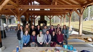 A group of women from TNC in Massachusetts pose for a photo on a picnic table under a wooden pavilion.