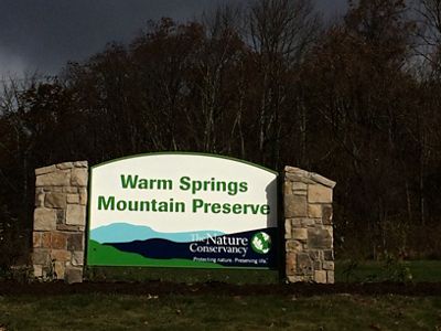 A large welcome sign reads "Warm Springs Mountain Preserve". The text is printed above a stylized graphic of two mountain ridges. TNC's name and globe logo is in the lower right corner.