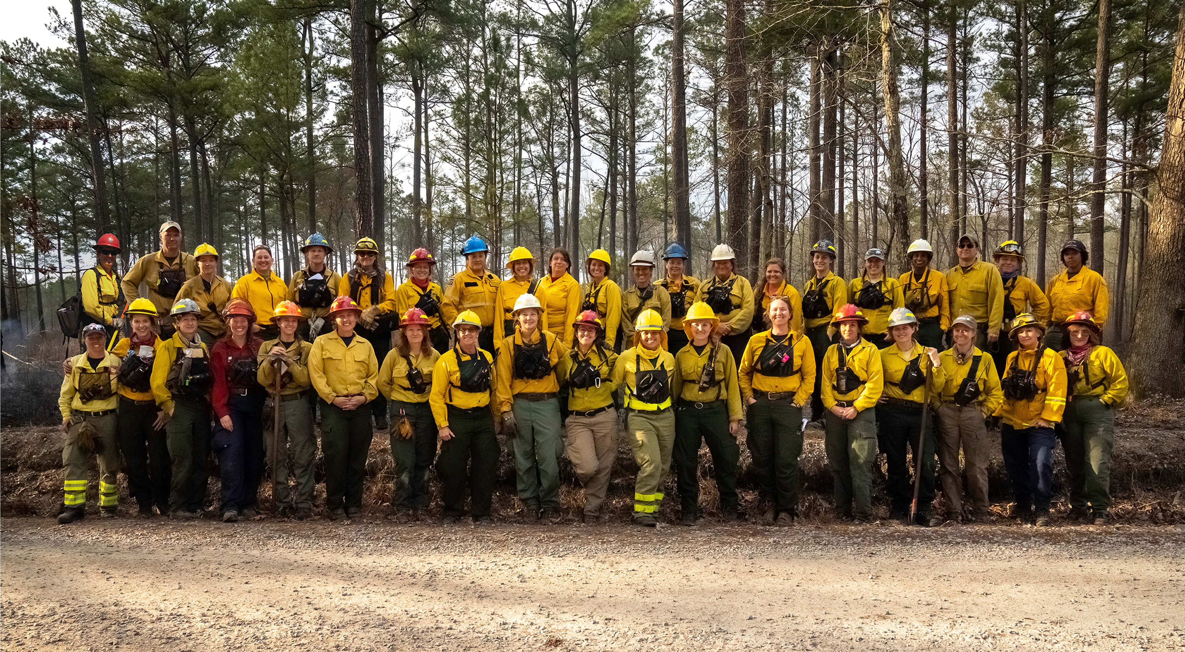 A large group of people pose together at the edge of a pine forest during a fire learning exchange.