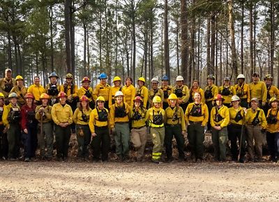 A large group of people dressed in fire gear pose together at the edge of a pine forest during a fire learning exchange.