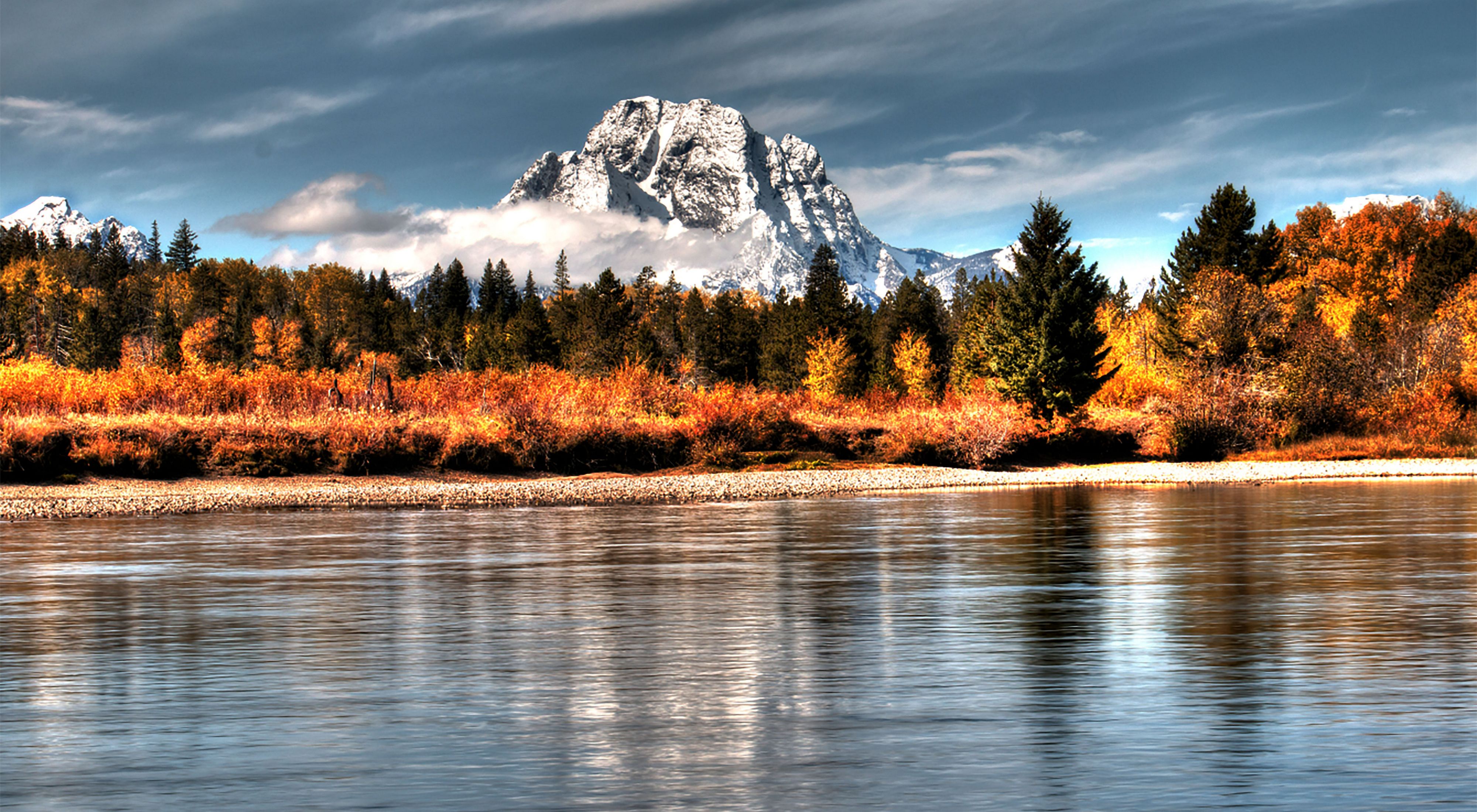 Bright orange fall foliage on the shores of Jackson Lake, with a large, rocky, snow-covered mountain peak in the background.