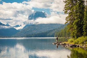 A man and child stand on the rocky shore of a wide lake. In the distance, forests and a mountain are shrouded in low clouds.