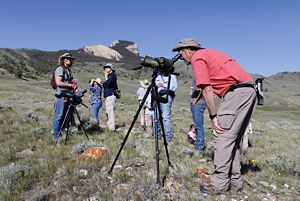 A man, surrounded by a group of bystanders, looks through scope in field.