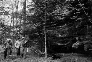 A black and white photo of three people standing in a forest next to a large rocky outcrop.