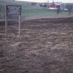 A faded color archival photo of a wooden sign marking Thousand's Rock Point, an Original Prairie stands in a prairie landscape, with rolling farmland, red barns, and a silo visible in the background.