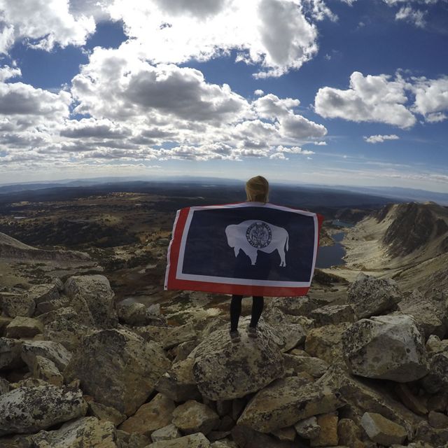 Woman facing away from the camera with arms outstretched holding the Montana flag while gazing across a valley from a rocky mountain peak.