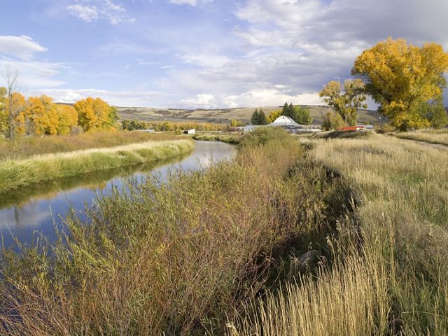 A river runs between grass covered banks, curving into the distance. A low farm building sits by the river at the curve. Low hills rise in the distance. 