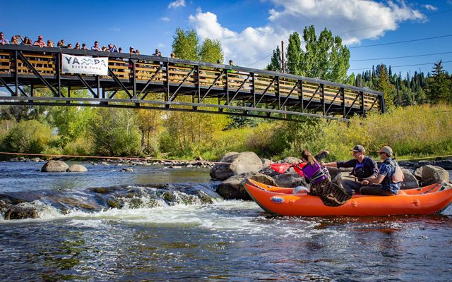 People in a boat cut a ribbon stretched across the Yampa River while spectators watch from a nearby bridge that extends across the river. 