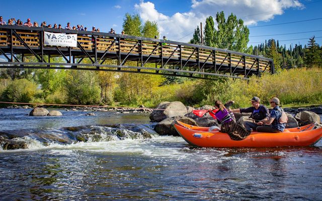 People in an orange inflatable boat on a river. A bridge with onlookers are above them.