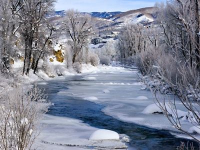 A winter landscape on the Yampa River showing a narrow portion of river flowing through its frozen edges, with bare, snow-covered trees along the banks and snowy mountains in the background. 