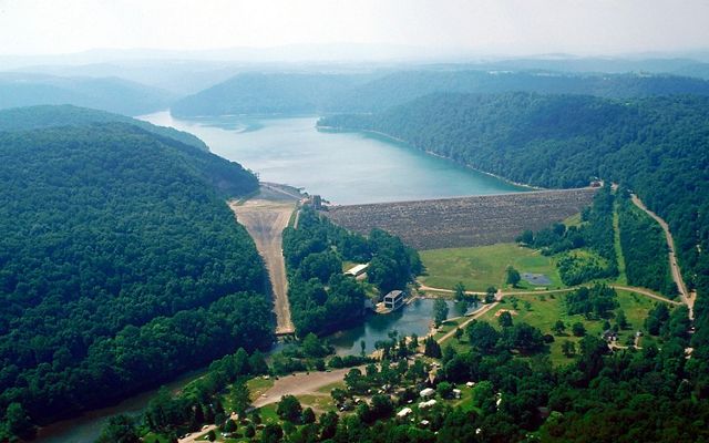 A large lake backs up behind a tall concrete dam. The dam spans a distance between two forest covered mountains. A small retaining pond sits in front of the dam.