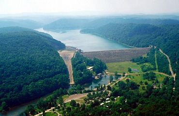 A large lake backs up behind a tall concrete dam. The dam spans a distance between two forest covered mountains. A small retaining pond sits in front of the dam.