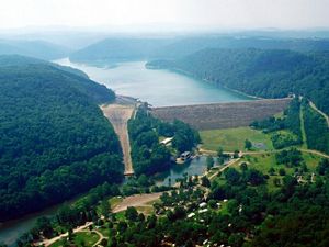 A large lake backs up behind a tall concrete dam. The dam spans a distance between two forest covered mountains. A small retaining pond sits in front of the dam.