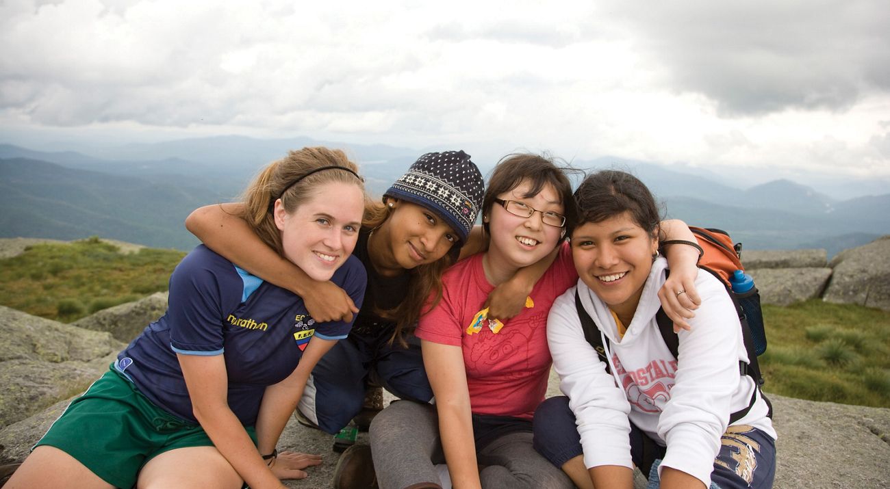 Four people sit together at a mountain overlook. There arms are linked around each others necks as they lean in and post together.