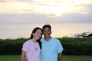 Eiichiro and Yumi Kuwana stand in a grassy area with the ocean behind them.