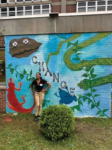 A young woman poses in front of a mural she painted that includes an oyster, salamander, snake and frog.