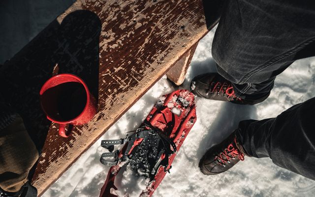 A view looking down at a person's feet in the snow next to a pair of snowshoes and a wooden bench with a red mug sitting on it.