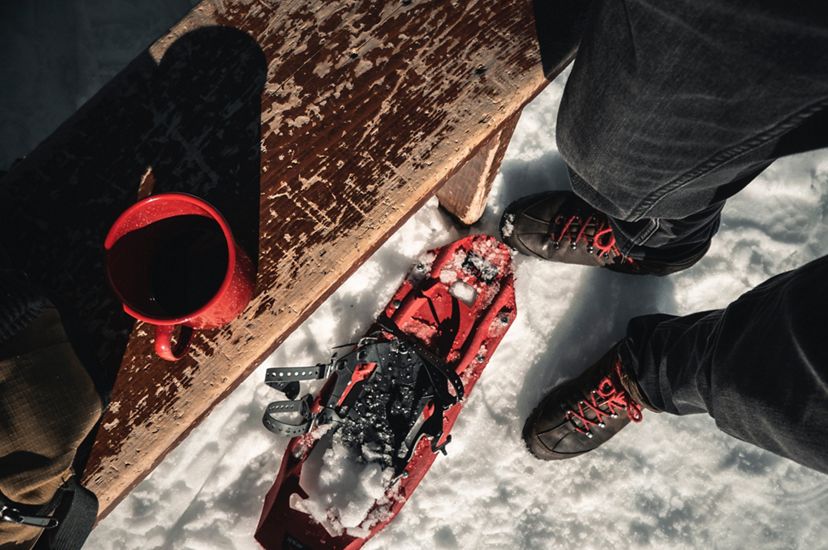 A view looking down at a person's feet in the snow next to a pair of snowshoes and a wooden bench with a red mug sitting on it.