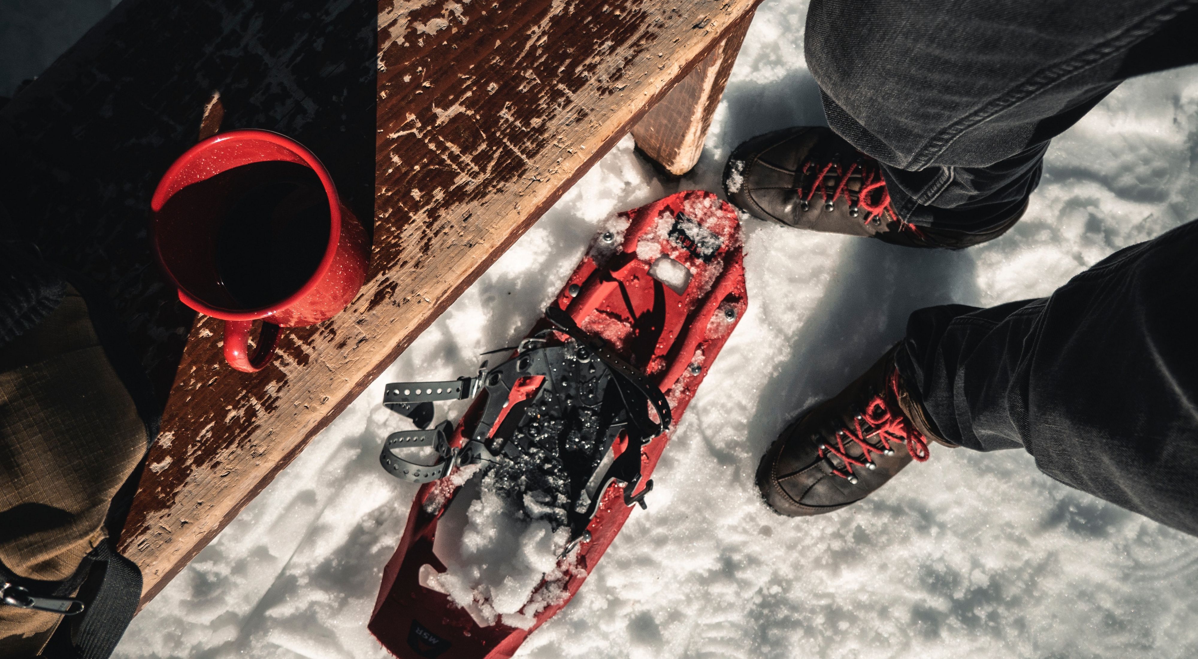 A view of a person's feet in the snow next to a pair of red snowshoes.