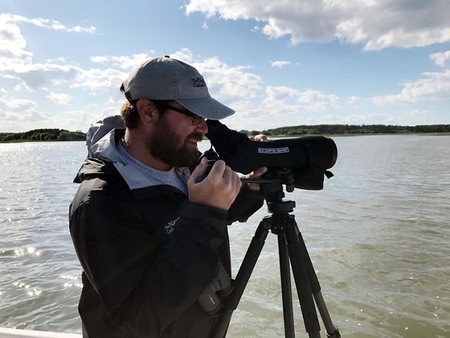 Candid snapshot of Coastal Biologist Zak Poulton. A bearded man looks through a monocular scope mounted on a tripod to observe birds at the Virginia Coast Reserve during TNC's 2017 Bird Blitz event.