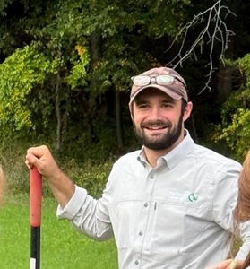 A man wearing a hat and Nature Conservancy hat sands with a shovel near a forest.