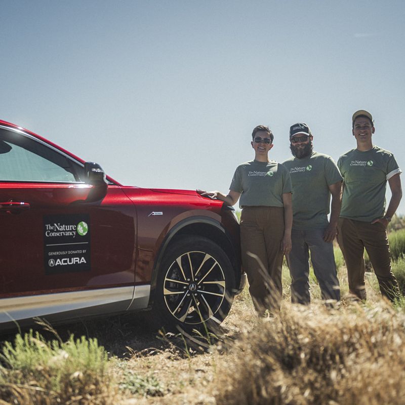 Three TNC employees stand next to an electric SUV on a grassy hill at TNC's Frank and Joan Randall preserve in the Tehachapi foothills.